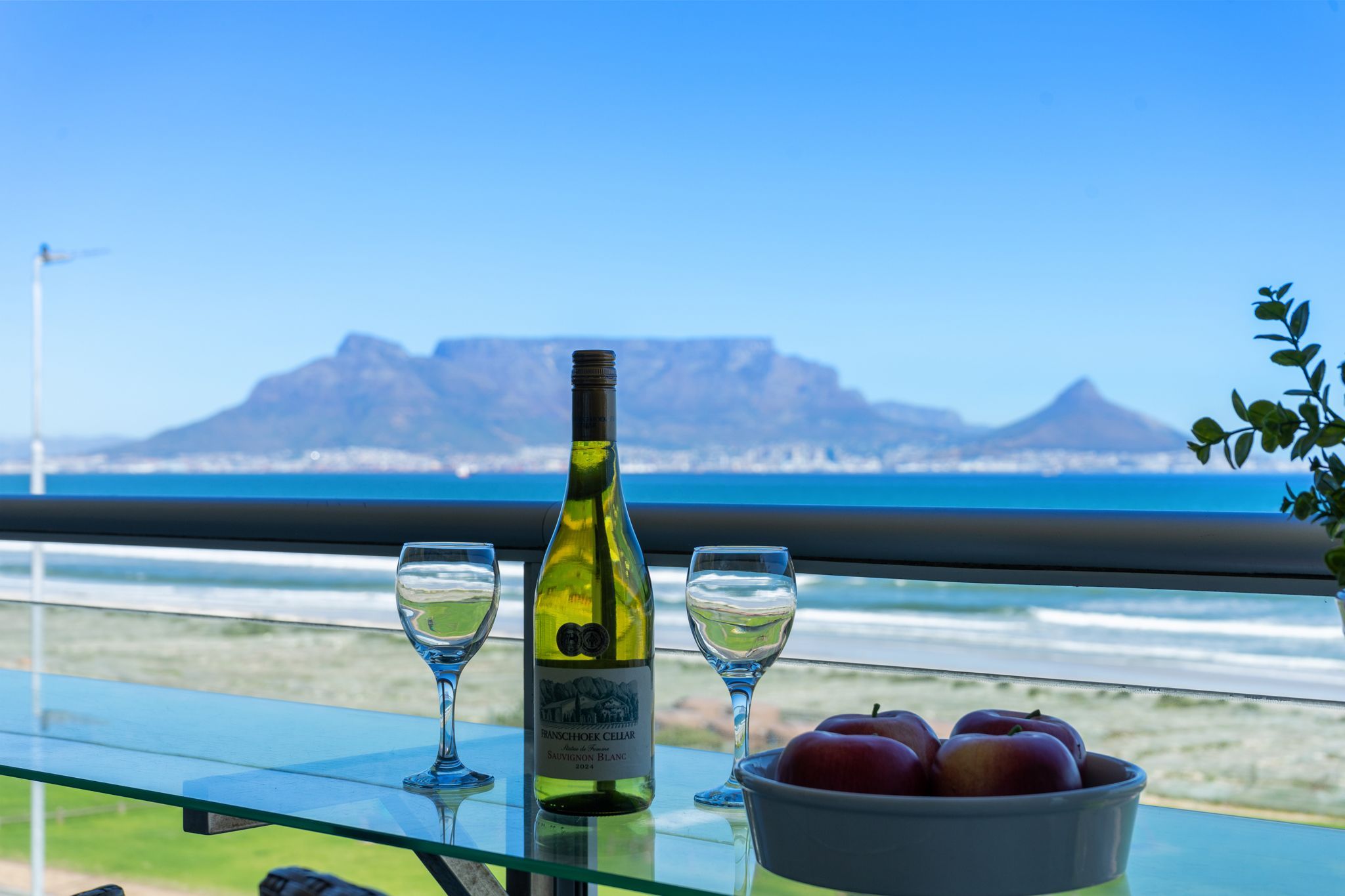 Eine Weinflasche, zwei Gläser und eine Schüssel Äpfel auf einem Tisch mit Blick auf Berge und Meer.
