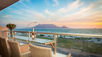 A balcony with wicker chairs and a glass table, overlooking a coastal view with a mountain in the distance.