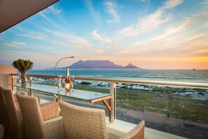 A balcony with wicker chairs and a glass table, overlooking a coastal view with a mountain in the distance. - Sea Spray B306 (Cape Town)