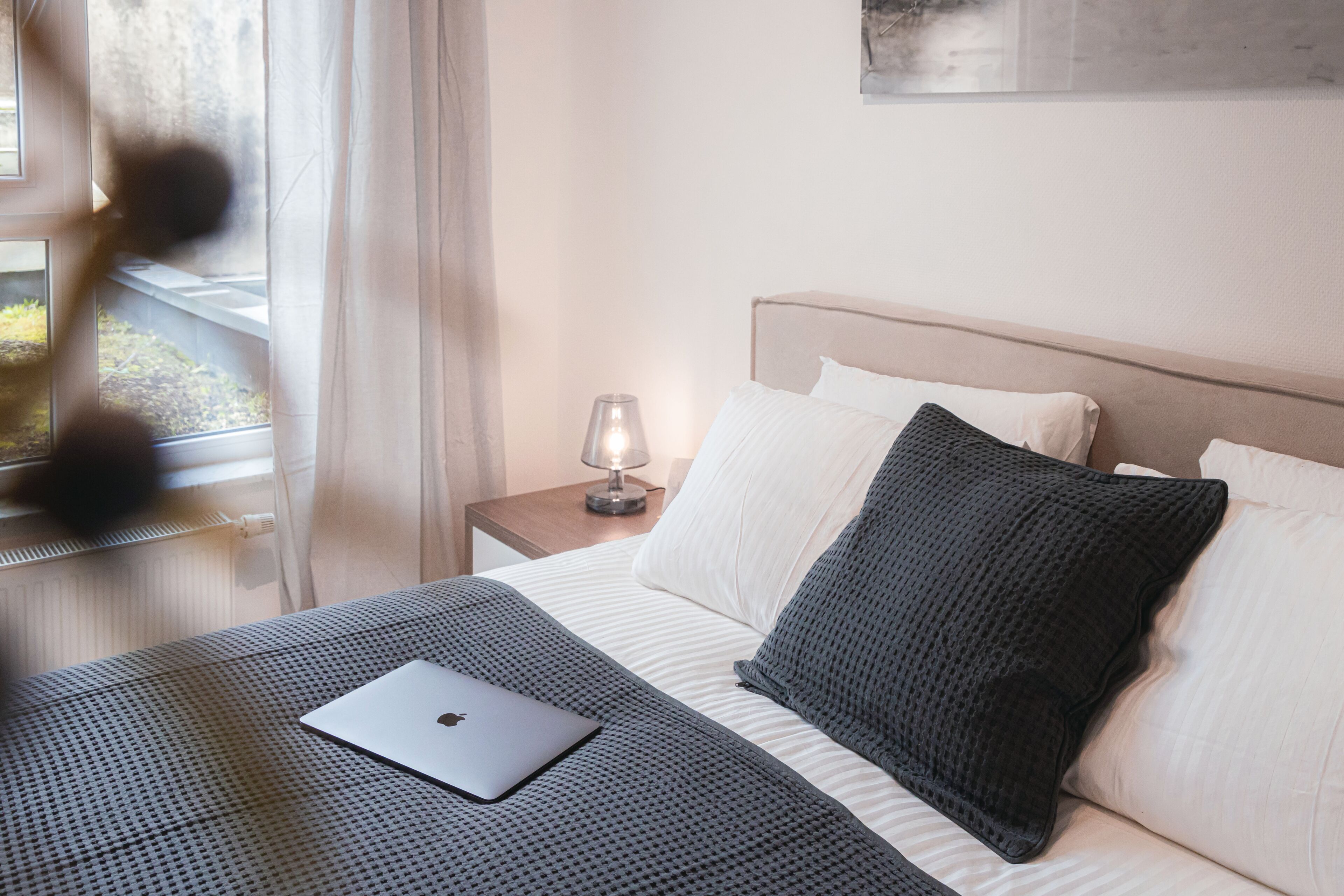 A neatly made bed with a laptop, a bedside lamp, and a window with curtains.