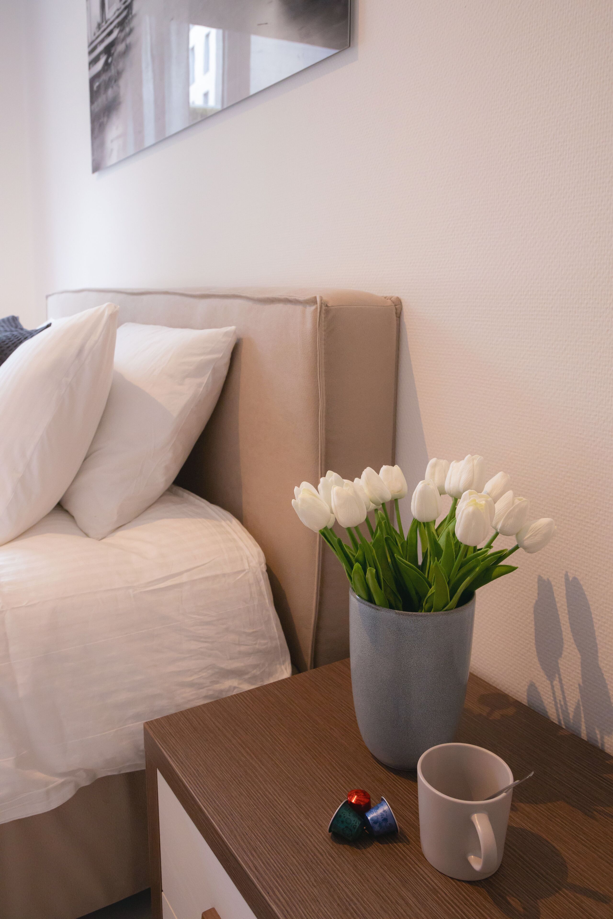 Ein Hotelzimmer mit einem Bett, einem Nachttisch, einer weißen Tasse und einer Vase mit weißen Tulpen.