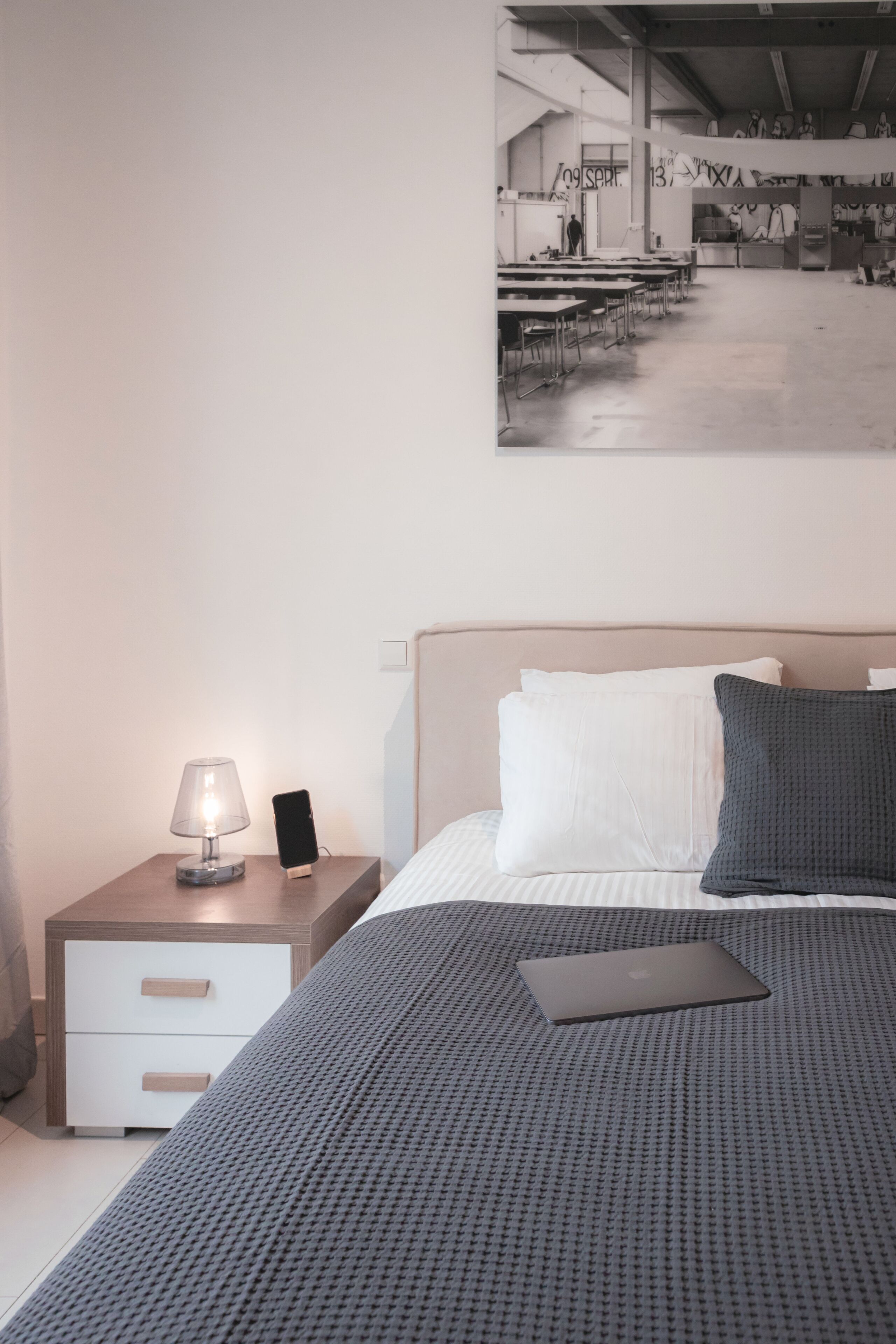 Une chambre à coucher bien rangée, avec un lit, une lampe de chevet, un ordinateur portable et une œuvre d’art accrochée au mur représentant une scène d’atelier.