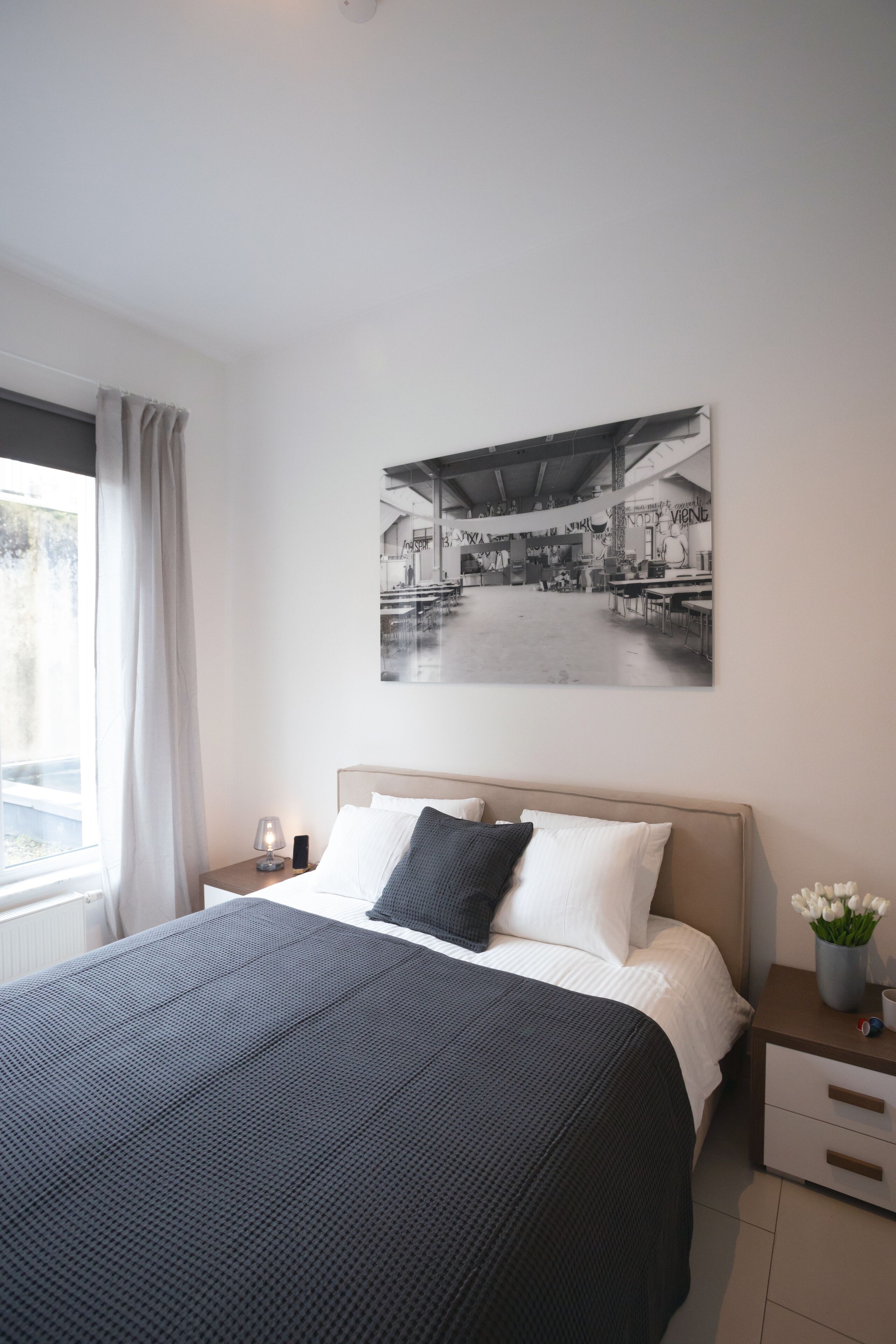 Camera da letto con un letto, un comodino, una lampada, un vaso con fiori e un quadro appeso al muro.