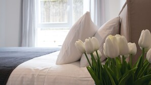 A bed with white bedding and pillows, a vase with white tulips, and a window with daylight.