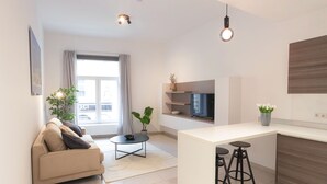 A modern living room with a sofa, a coffee table, and a TV. There is a kitchen area with a counter and stools.