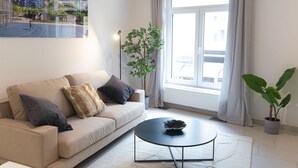 A modern living room with a beige sofa, a black coffee table, a large window with curtains, and a potted plant.