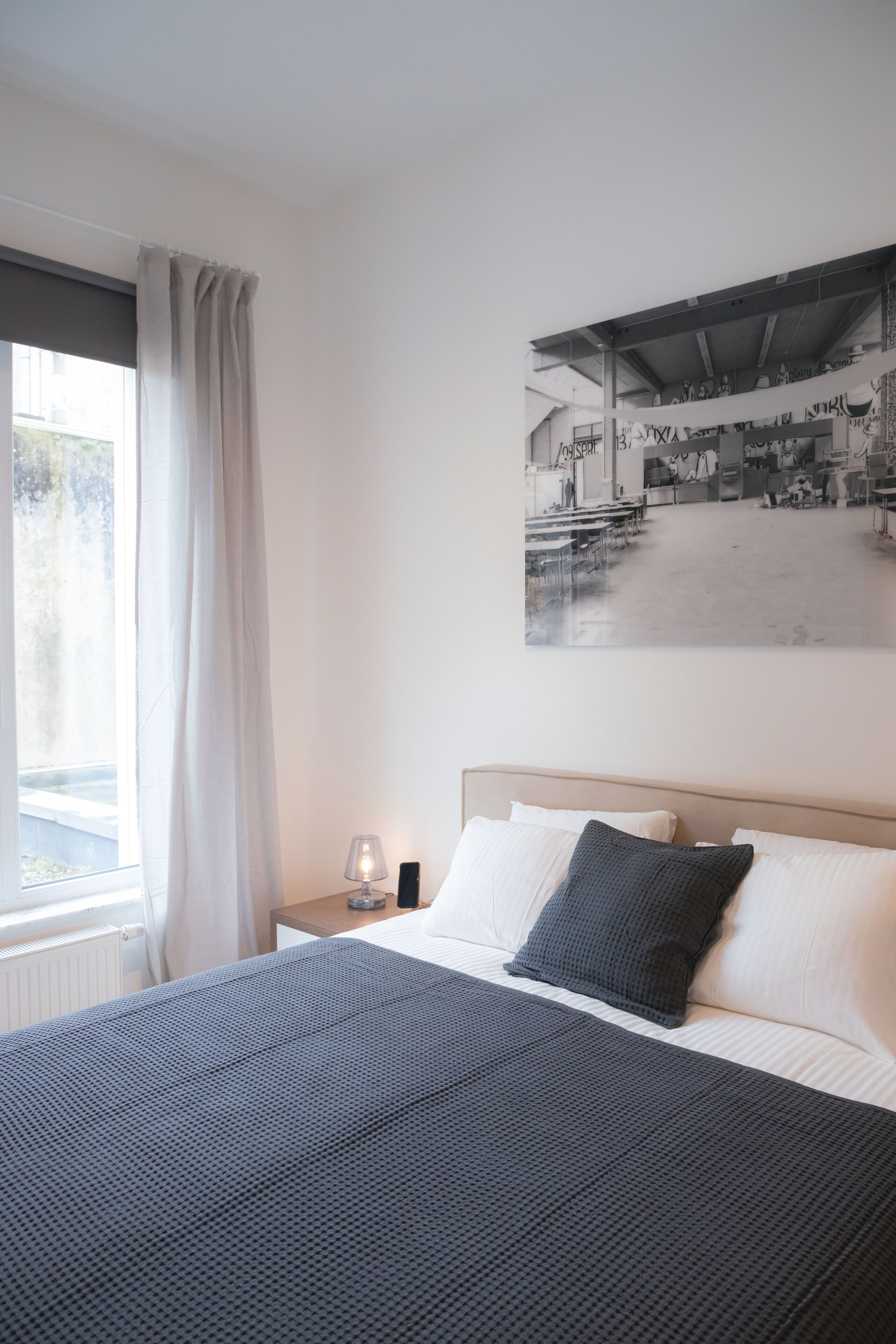 Un dormitorio con una cama grande, una mesita de noche con lámpara, una ventana con cortinas y un cuadro colgado en la pared.