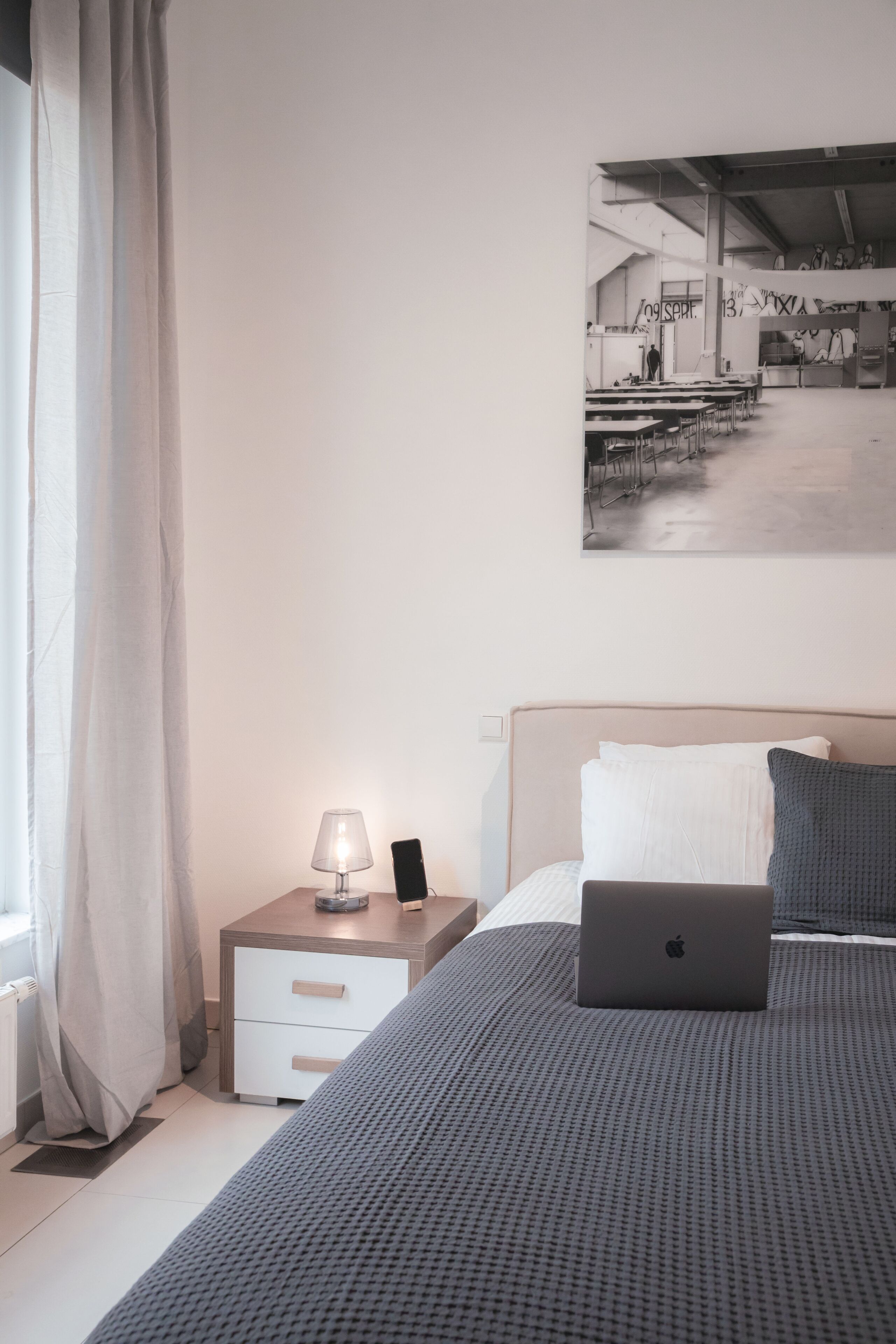 A bedroom with a bed, a nightstand with a lamp, a laptop, and a painting on the wall.
