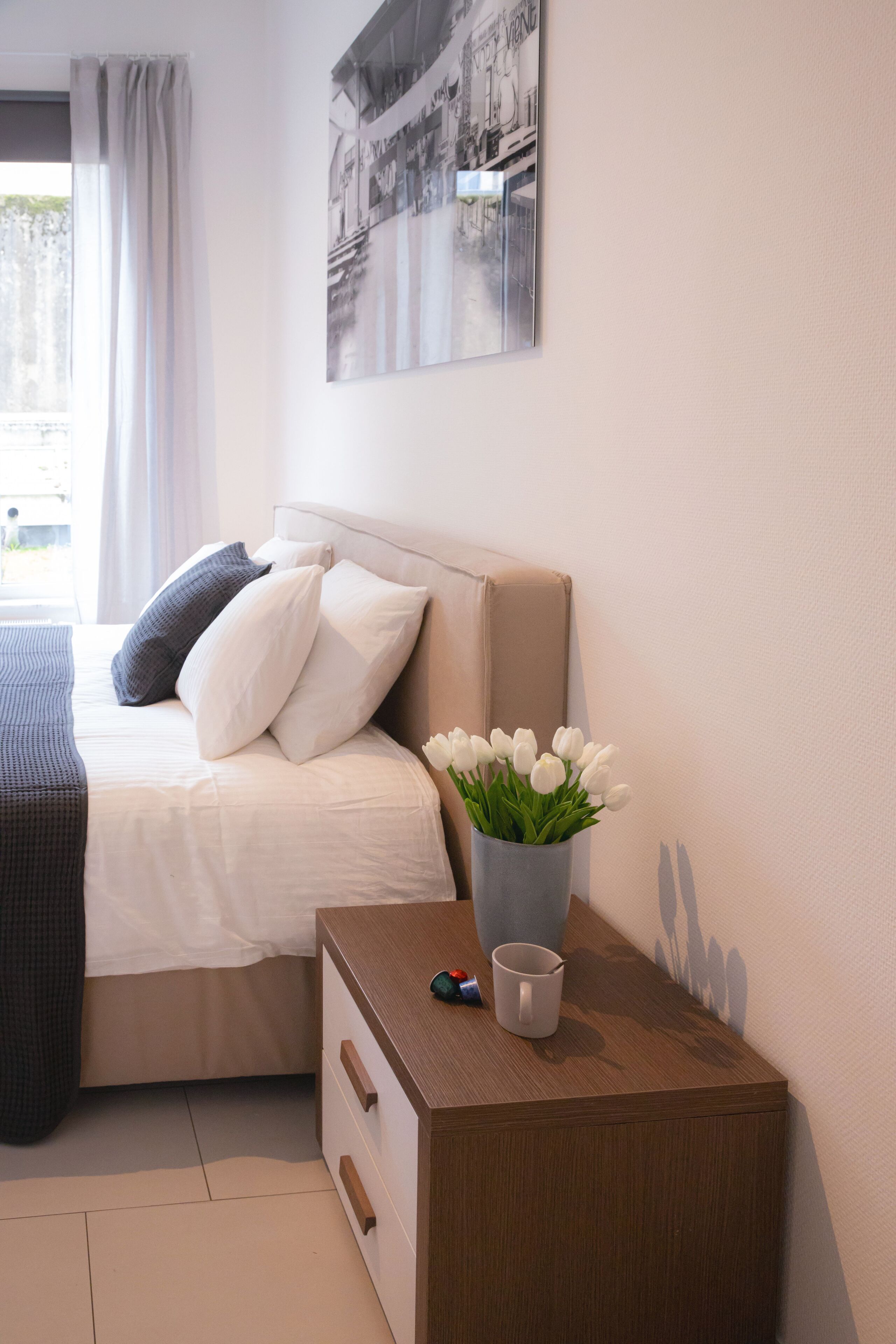 A neatly arranged bedroom with a bed, a bedside table with a vase of flowers, and a wall-mounted picture.