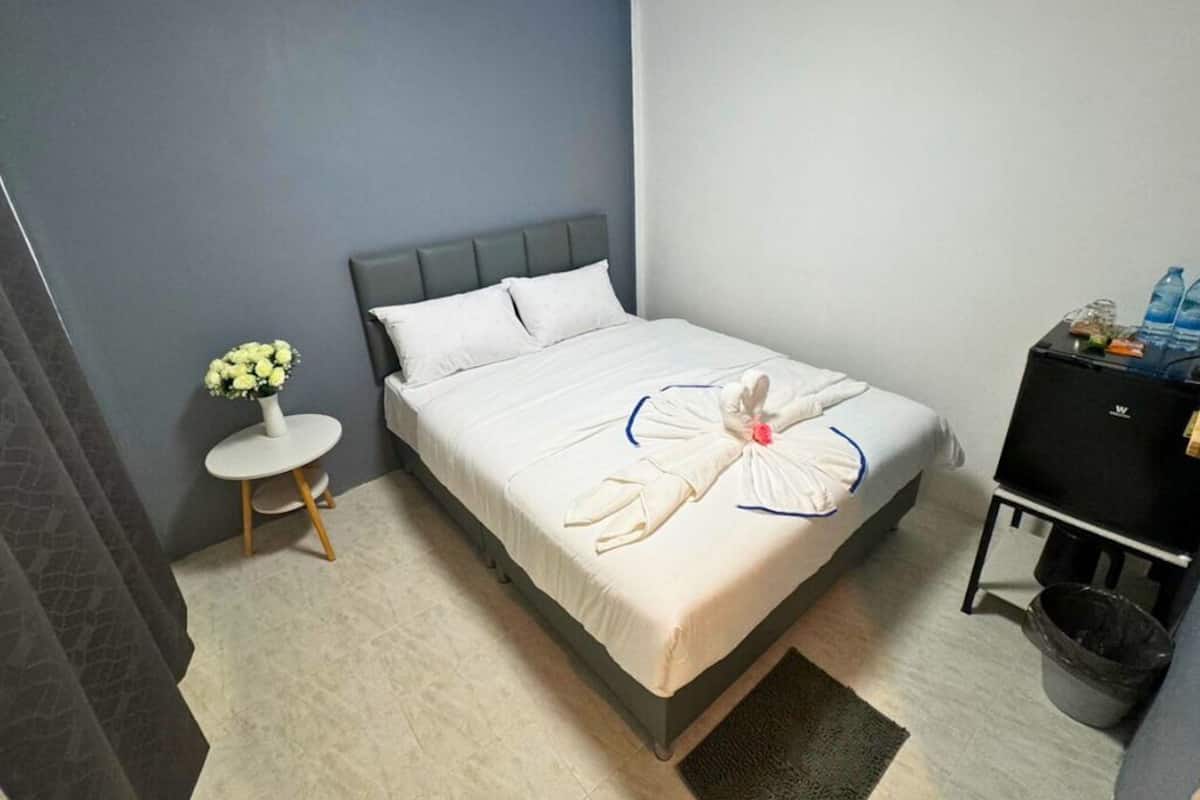 A neatly made bed with a towel art design, a small round table with a vase of flowers, and a black cabinet with items on it.