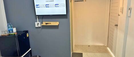 A hotel room with a bed, a television mounted on the wall, a small desk with a computer, and a door leading to a shower area.