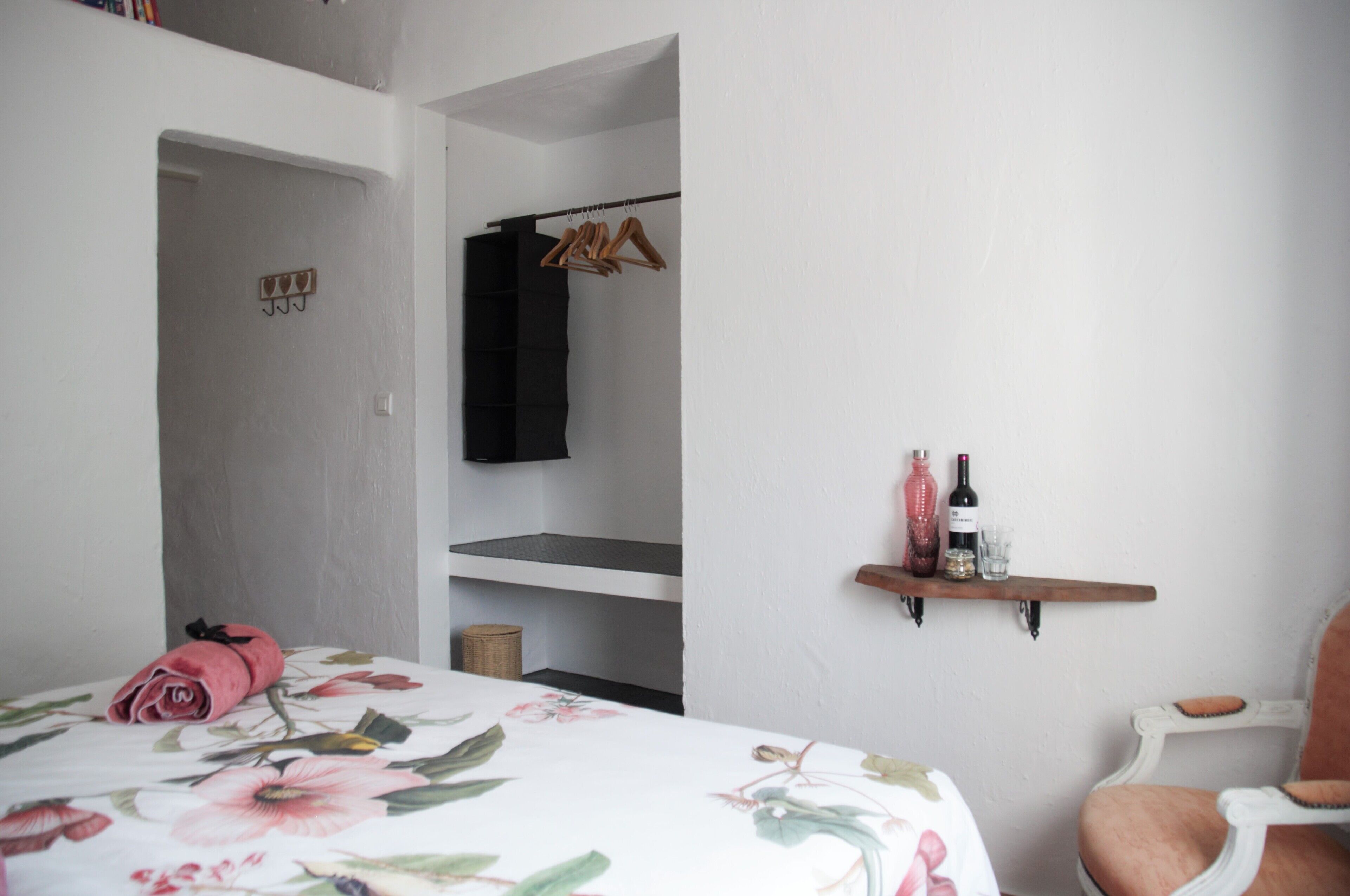 Un dormitorio con una colcha estampada, una toalla enrollada sobre la cama, un perchero, una estantería con botellas y vasos, y una silla.