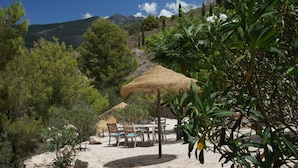 A rustic outdoor seating area with a thatched umbrella, surrounded by greenery and a mountainous backdrop. - Boutique B&B El Refugio (Alcaucin)