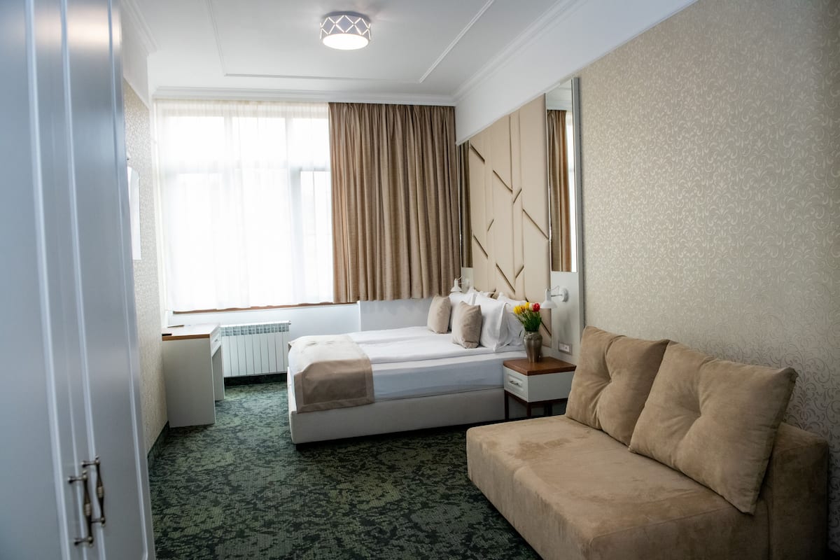 Quarto de hotel com cama, sofá, mesa de cabeceira, espelho e janela com cortinas.