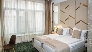 Una habitación de hotel con una cama grande, una silla, una mesita de noche con lámpara y una ventana con cortinas translúcidas.