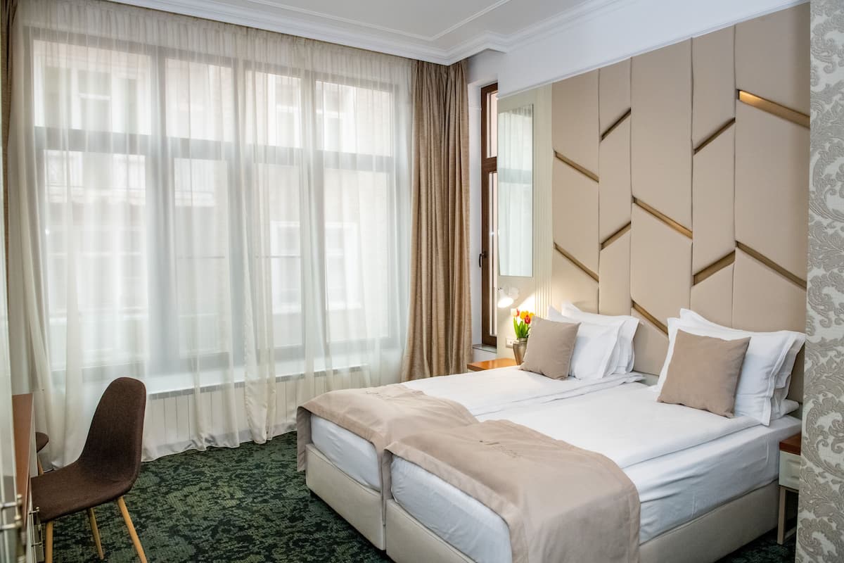 Quarto de hotel com uma cama grande, uma cadeira, uma mesinha de cabeceira com abajur e uma janela com cortinas leves.
