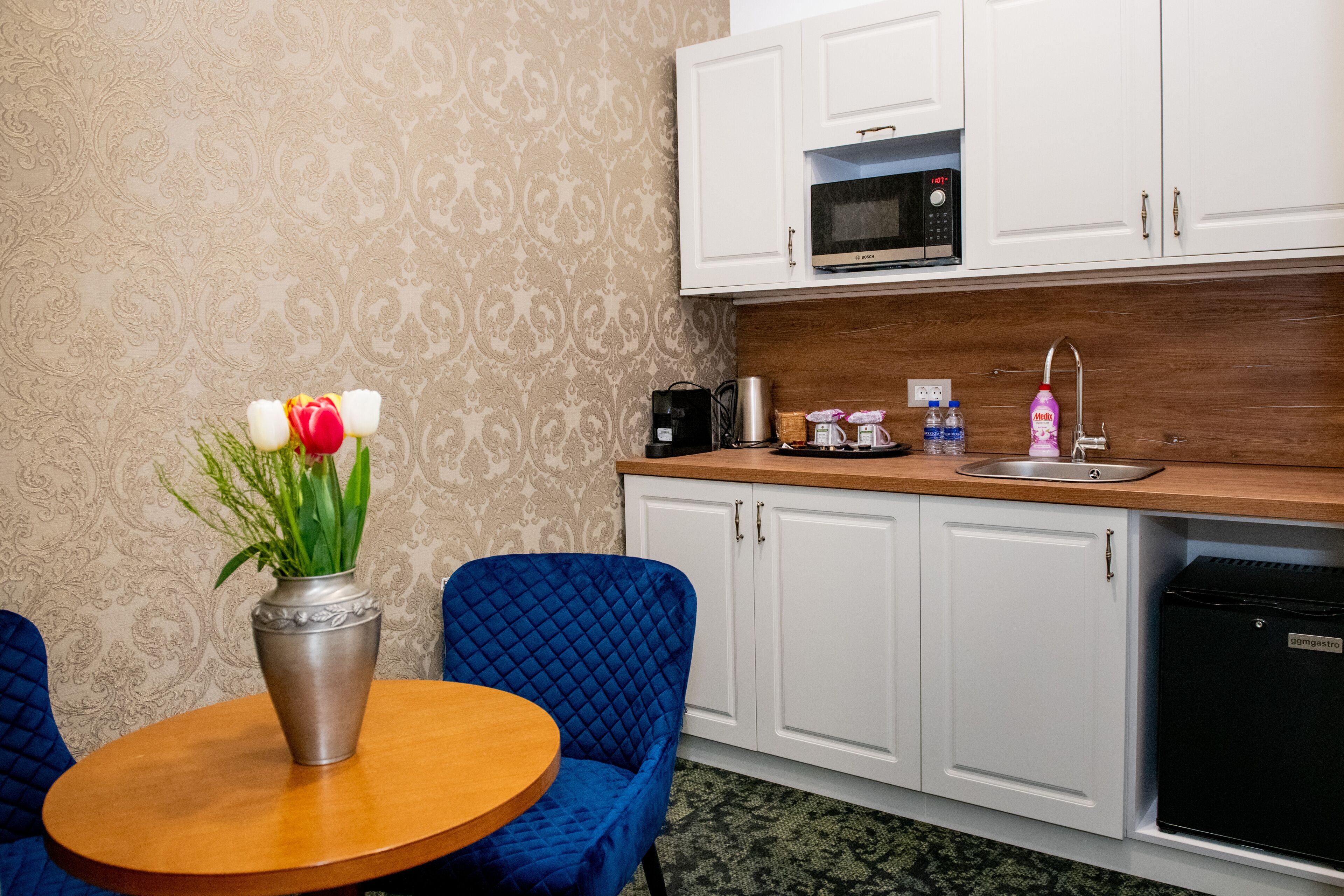 Cocina compacta con armarios blancos, microondas, lavabo y refrigerador. Hay una mesa redonda de madera con un florero y dos sillas tapizadas en azul.