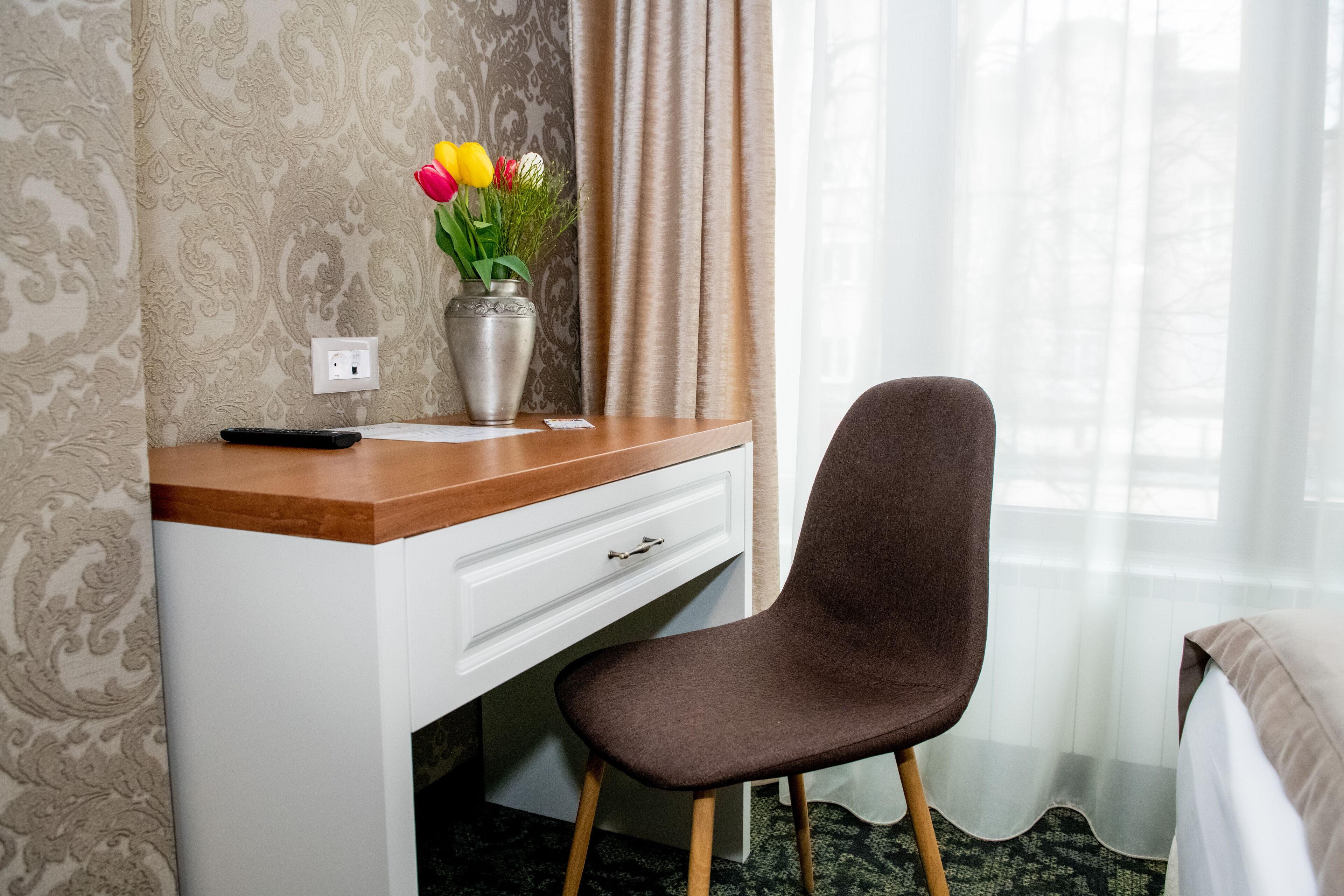 A hotel room with a wooden desk, a brown chair, a vase with flowers, and a bed.