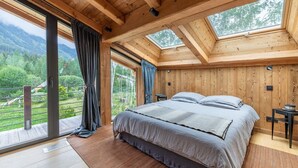 Ein Schlafzimmer mit einem großen Bett, Holzbalkendecke und Blick auf eine Berglandschaft durch große Fenster.