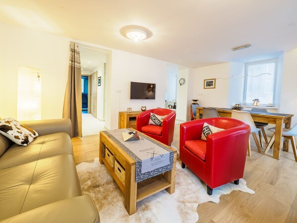 A modern living room with a sofa, two red armchairs, a wooden coffee table, and a TV mounted on the wall.
