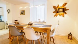 Uma sala de jantar com mesa de madeira, quatro cadeiras, um objeto decorativo na parede e uma janela com luz natural.