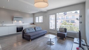 Una sala de estar moderna con un sofá gris, una mesita y un televisor. Hay una zona de cocina con armarios blancos y un horno empotrado. Un ventanal ofrece vistas a edificios exteriores.