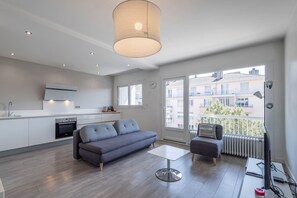 A modern living room with a grey sofa, a small table, and a TV. There is a kitchen area with white cabinets and a built-in oven. A large window offers a view of buildings outside. - SmartStay Le Thiou - Annecy centre (Annecy)