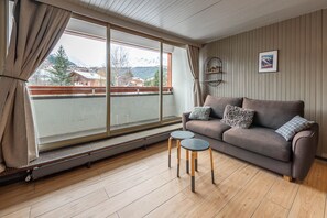 Ein gemütliches Wohnzimmer mit einer Couch, zwei kleinen Tischen, einer Heizung und einem großen Fenster mit Vorhängen.
