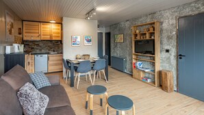 A modern kitchen with wooden cabinets, a dining area with a table and chairs, a living room with a sofa and two stools, and a bookshelf with various items. - SmartStay Grand Soleil - Courchevel 1650 (Courchevel)