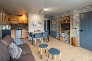 Eine moderne Küche mit Holzausstattung, ein Essbereich mit Tisch und Stühlen, ein Wohnzimmer mit Sofa und zwei Hocker sowie ein Bücherregal mit diversen Gegenständen.