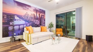 A modern living room with a white sofa, orange cushions, a glass coffee table, and a cityscape mural on the wall. - Luxury Living Steps from The Domain: Austin's Premier Shopping Haven! (Austin)