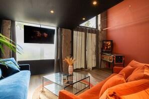 Un salon moderne avec un canapé bleu, un canapé orange, une table basse en verre et un téléviseur fixé au mur.