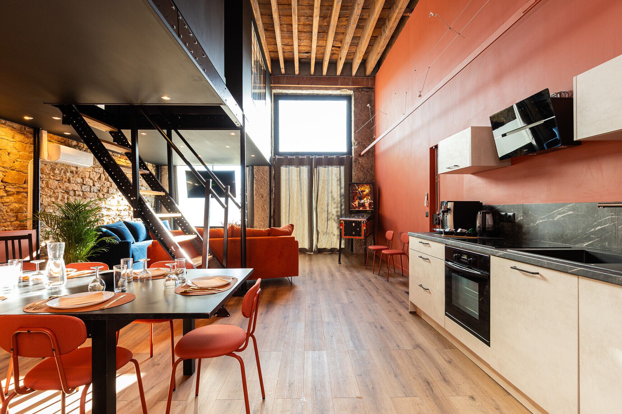 A modern kitchen with a wooden ceiling, a dining area with orange chairs, and a stone wall.