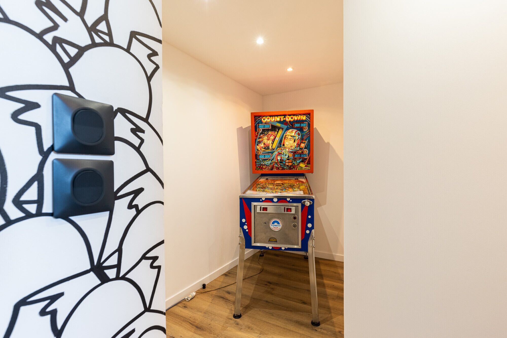 Un pasillo estrecho con una máquina de pinball frente a una pared con un diseño abstracto en blanco y negro.
