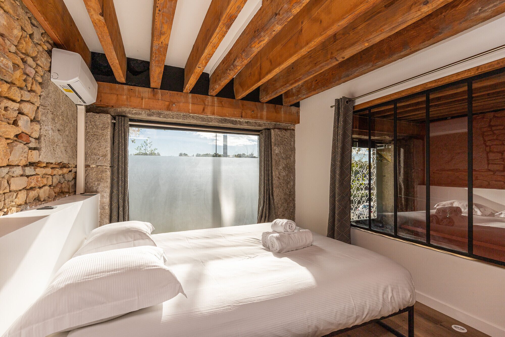Une chambre avec un mur en pierre, des poutres en bois, une grande fenêtre et un lit recouvert de draps blancs.