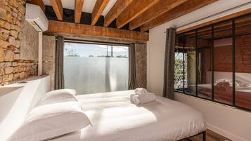 Une chambre avec un mur en pierre, des poutres en bois, une grande fenêtre et un lit recouvert de draps blancs.