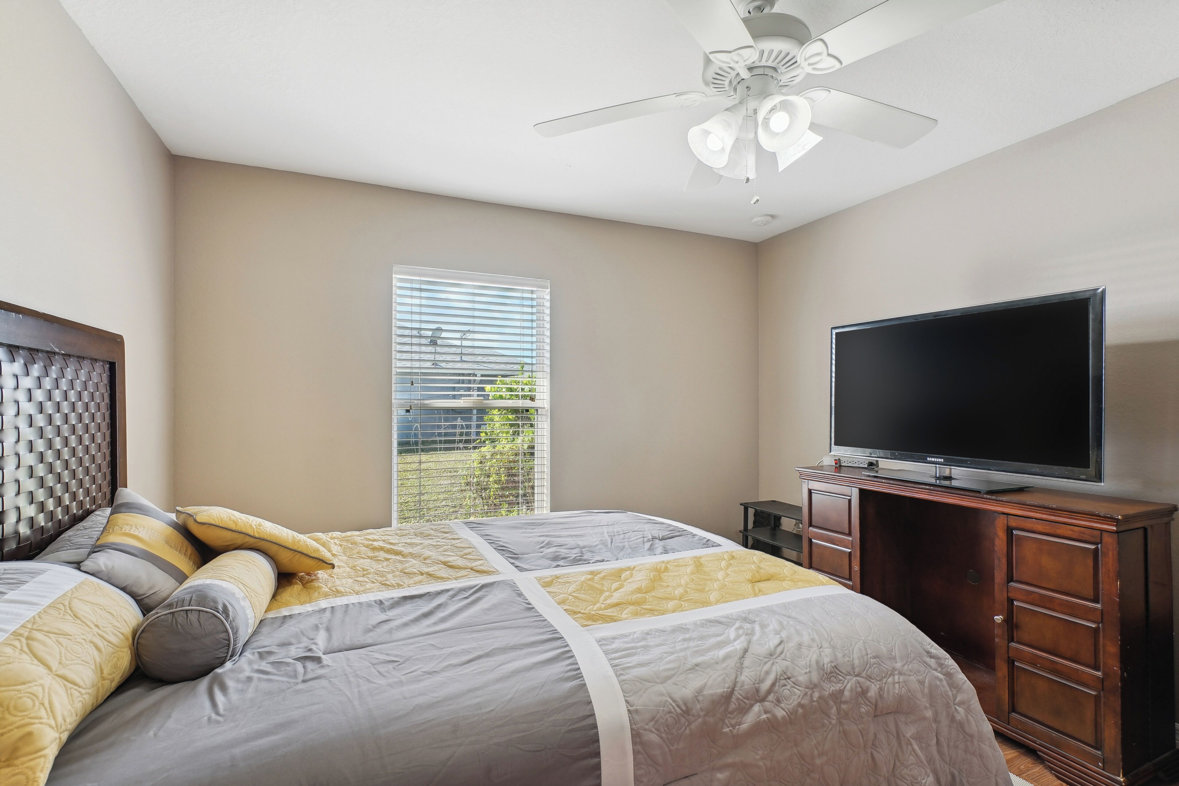 Un dormitorio con cama, televisión, ventilador de techo y ventana con persianas.