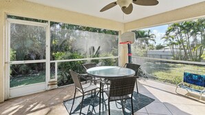 Une terrasse couverte avec une table en verre et des chaises en osier, un ventilateur de plafond et une vue sur les palmiers.