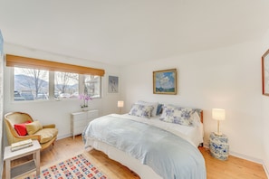 A bedroom with a bed, a chair, a nightstand, a lamp, a painting, and a window with a view of cars and mountains.