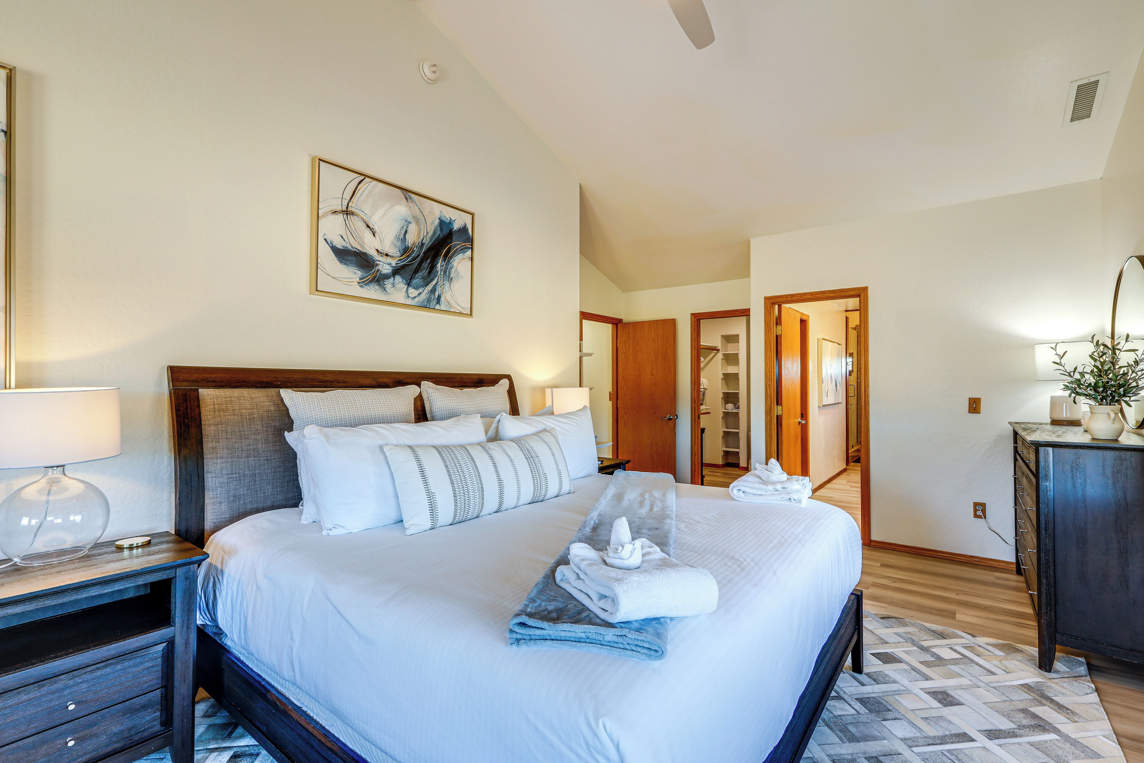 A hotel room with a large bed, a wooden headboard, a painting on the wall, a desk with a lamp, and a dresser.