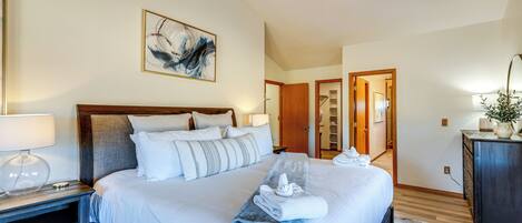A hotel room with a large bed, a wooden headboard, a painting on the wall, a desk with a lamp, and a dresser.