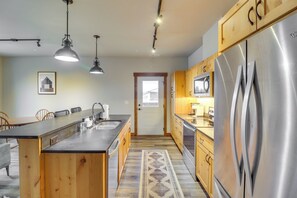 A modern kitchen with a central island, stainless steel appliances, wooden cabinets, pendant lighting, and a patterned rug. - Downtown Columbia Falls Condo: 16 Mi to Ski Resort (Columbia Falls)