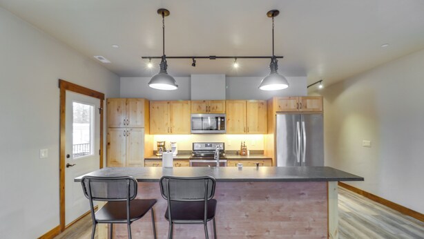 A modern kitchen with wooden cabinets, stainless steel appliances, and two bar stools. - Downtown Columbia Falls Condo: 16 Mi to Ski Resort (Columbia Falls)
