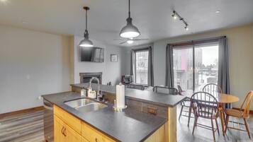 Una cocina con una isla central, un comedor con sillas, un ventilador de techo y un amplio ventanal con cortinas.