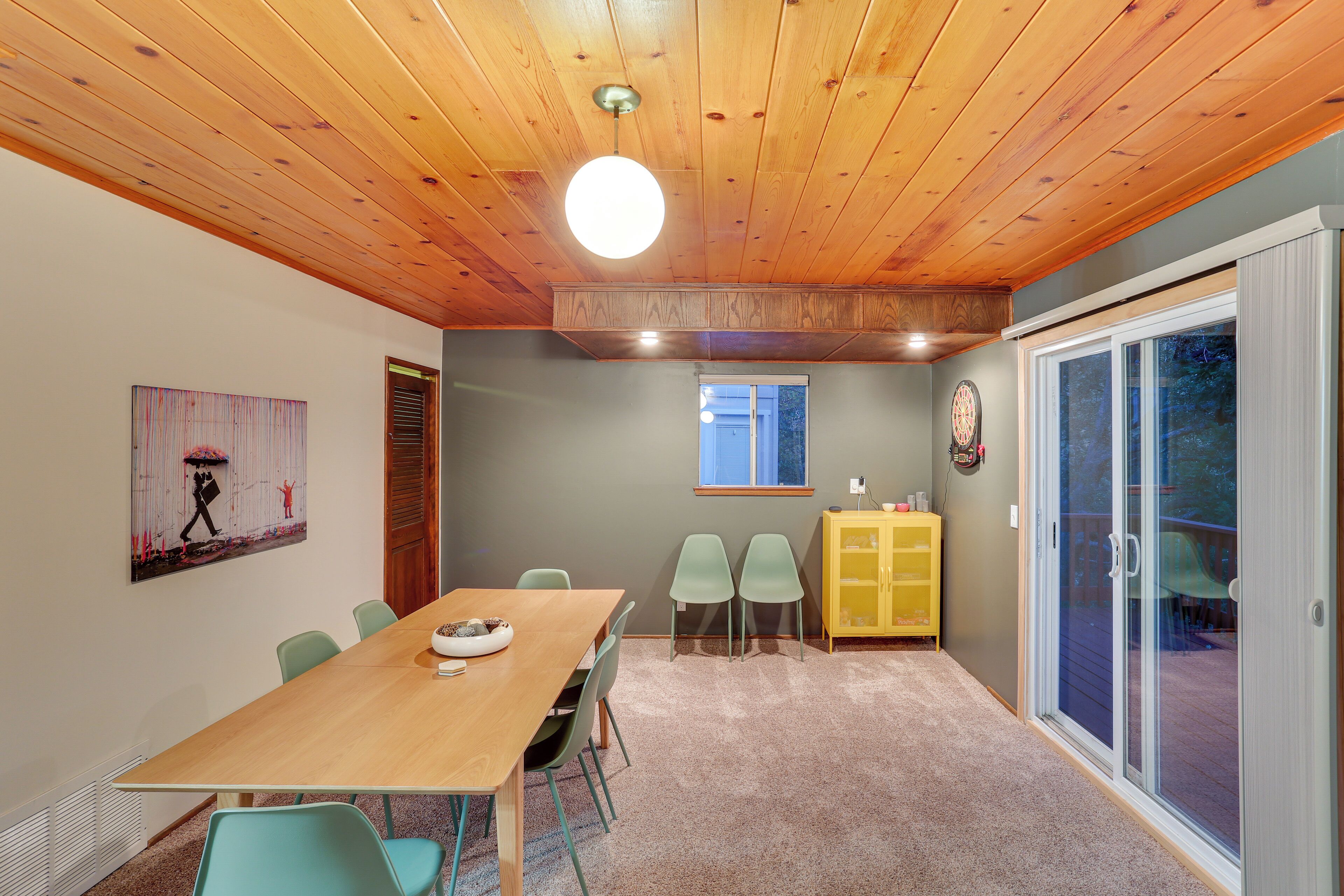 A room with a wooden ceiling, a dining table with chairs, a yellow cabinet, and a sliding glass door leading to an outdoor area.