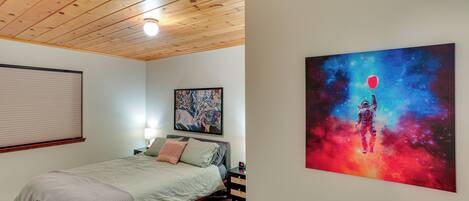 A bedroom with a wooden ceiling, a bed with a green bedspread, a nightstand with a lamp, and a painting of an astronaut on a red balloon.