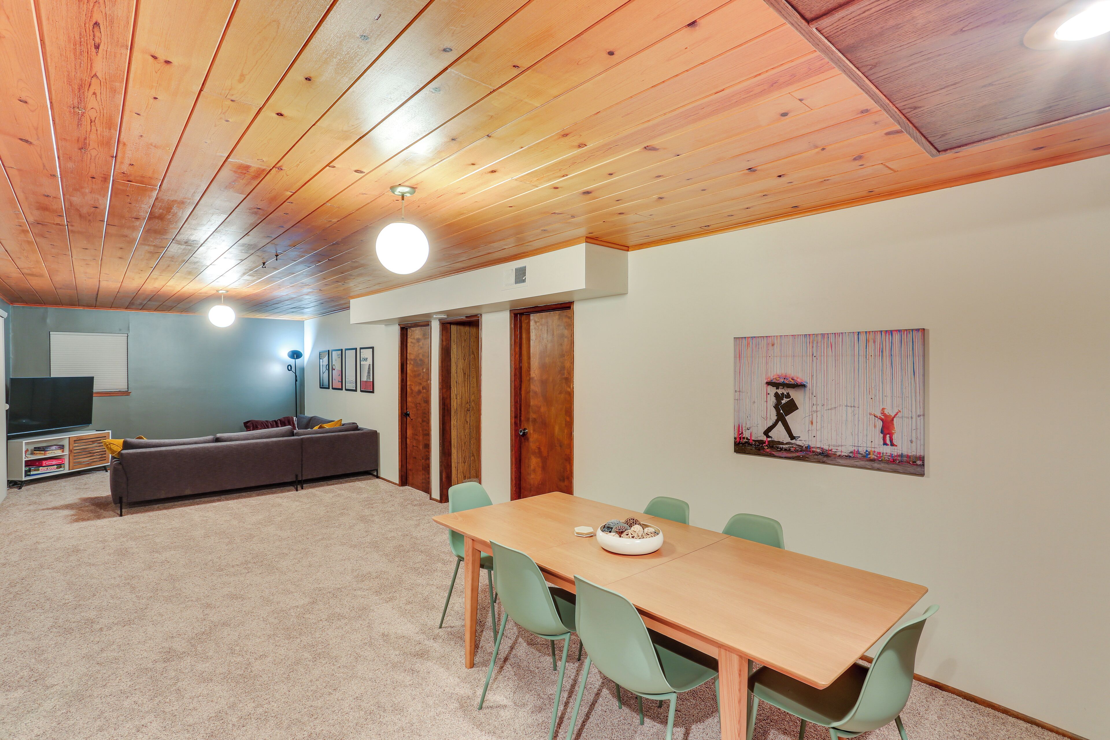 A room with a wooden ceiling, a dining table with chairs, a sofa, and a television.