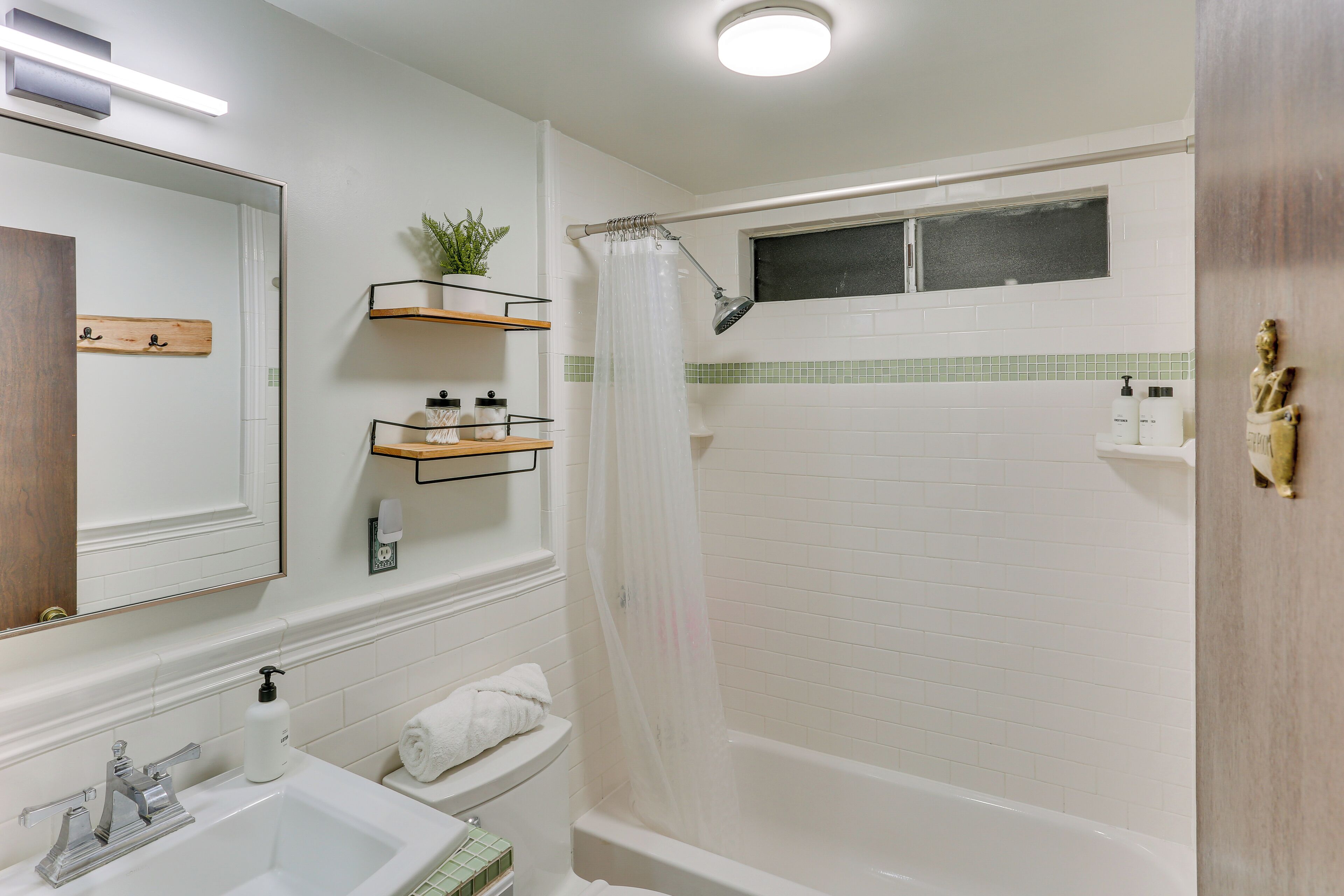 A modern bathroom with a white sink, mirror, and shower. There are shelves with toiletries and a potted plant.