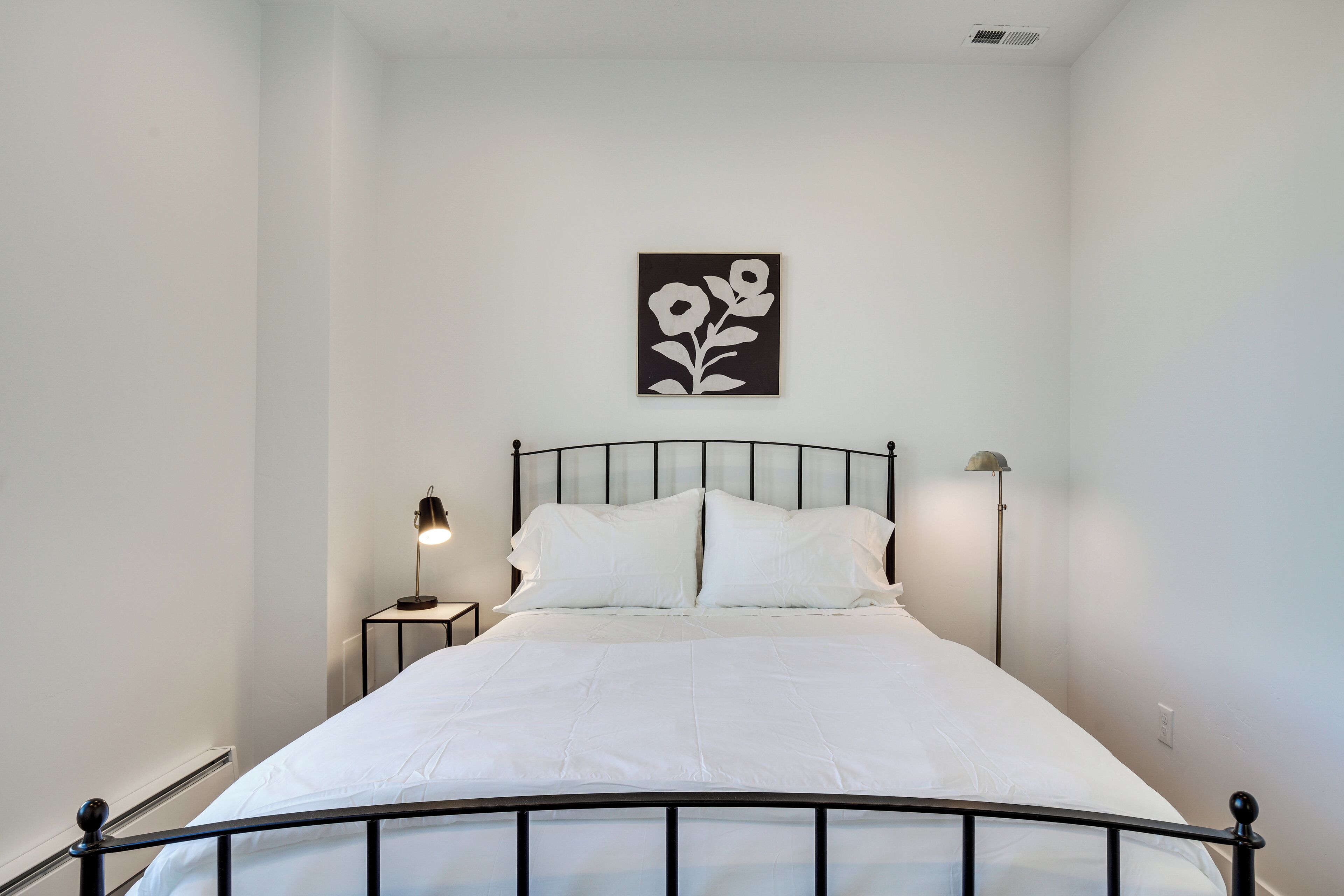 A neatly made bed with white linens, a black metal headboard, and a wall-mounted picture of flowers.