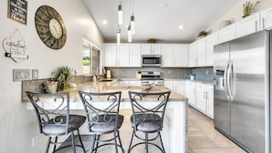 A modern kitchen with white cabinets, a stainless steel refrigerator, a large island with bar stools, and pendant lighting. - Gold Canyon Superstition Foothills Retreat w/ Pool (Gold Canyon)